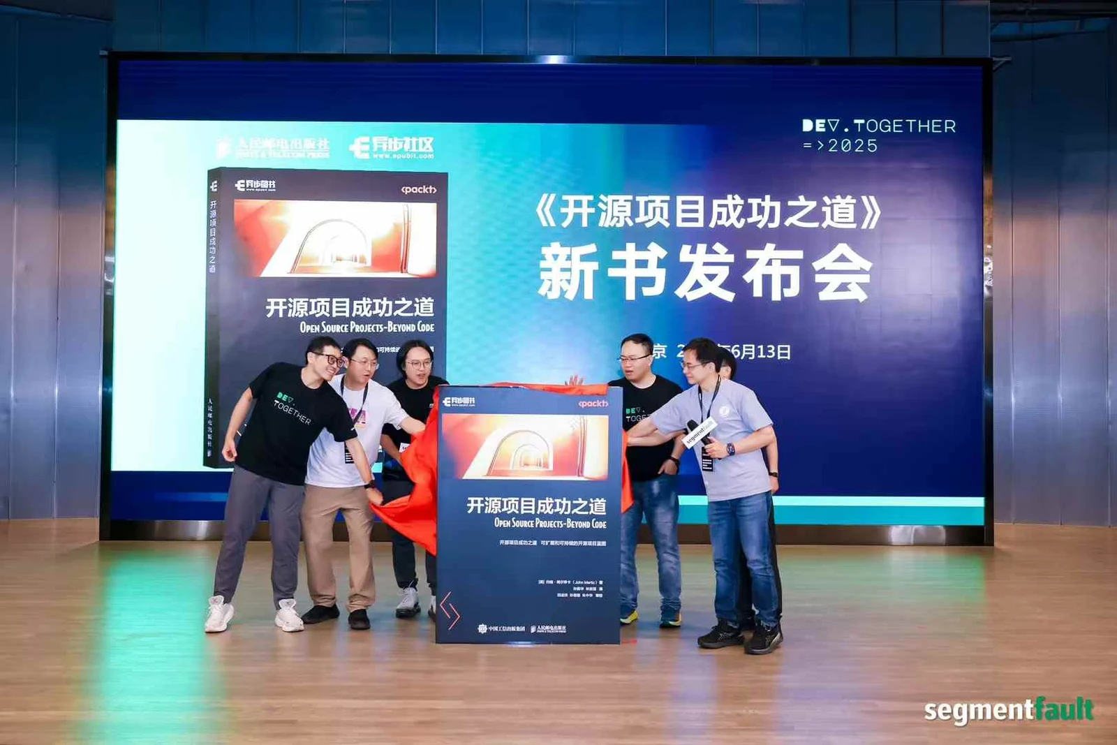 Five people on stage pulling a red cloth off a standee of Open Source Projects – Beyond Code at its Chinese launch in June 2025, with the book cover projected on the screen behind them.