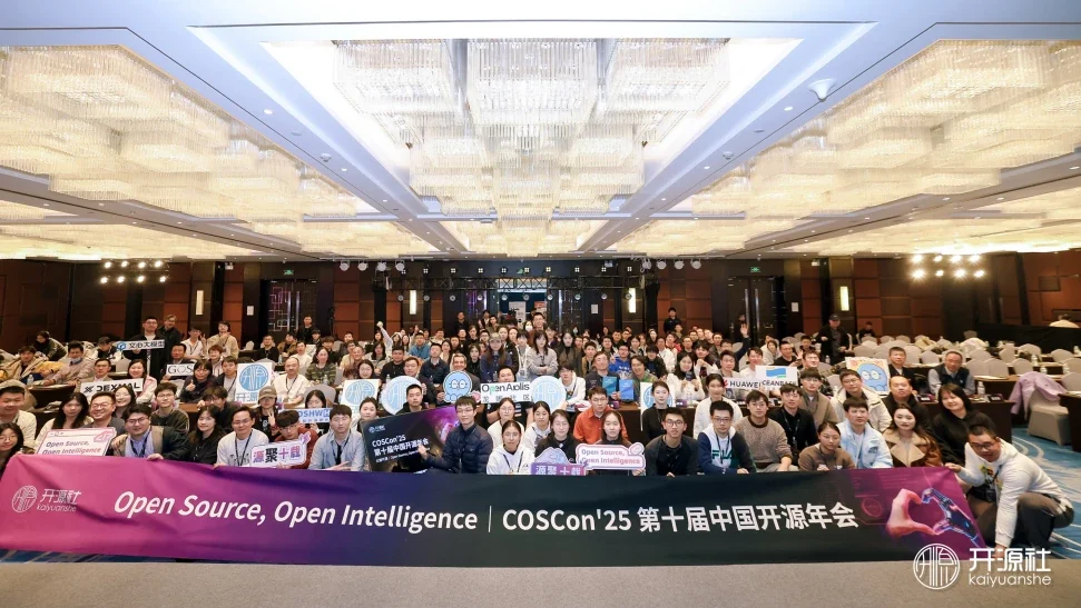A large group photograph of roughly a hundred and fifty attendees in a hotel ballroom, holding signs and standing behind a banner reading Open Source, Open Intelligence — COSCon'25.