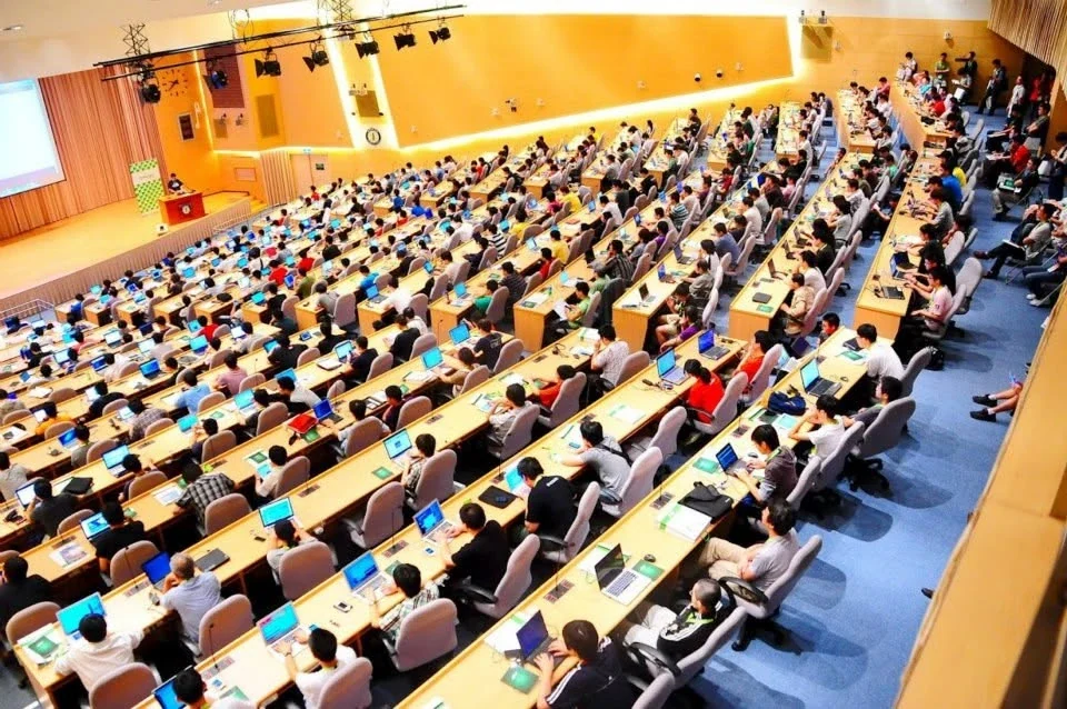 A packed tiered lecture hall seen from the back, every desk occupied and almost every attendee working on an open laptop, facing a speaker at a lectern beside a projection screen.